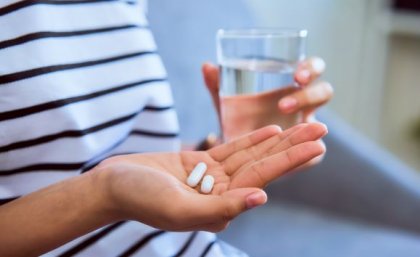 close up of a person's hand holding two small white capsules in the background is their other hand holding a glass of water